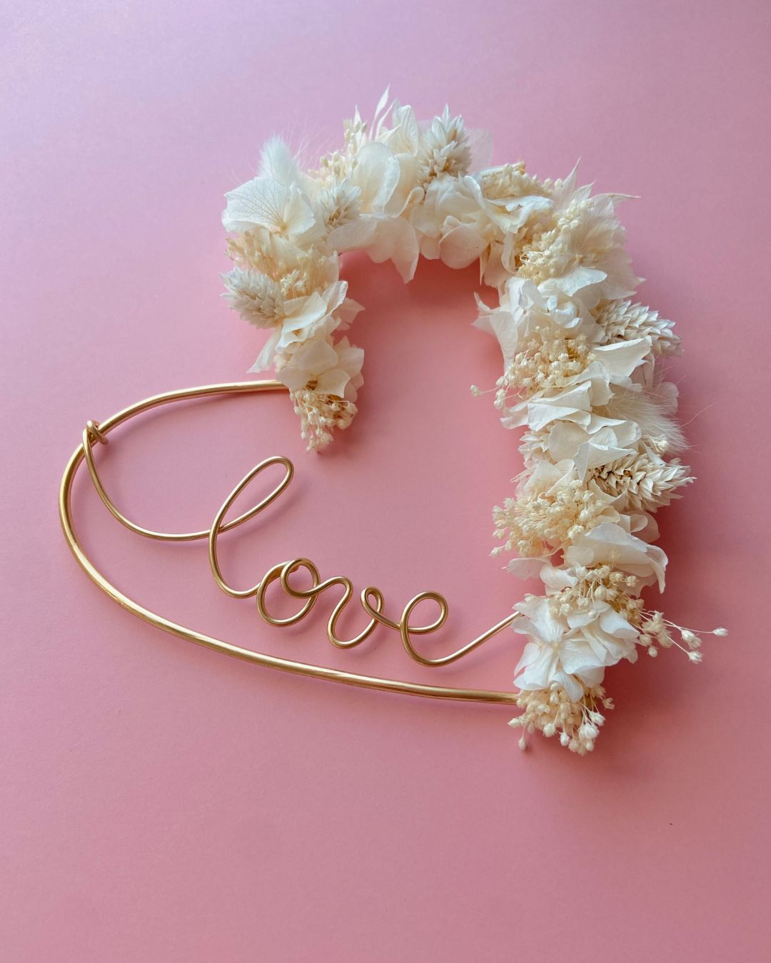 couronne en forme de coeur en fil doré avec inscription à l'intérieur LOVE en fil doré et fleurs séchées blanches sur le contour pour une décoration bohème chic