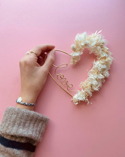 couronne en forme de coeur en fil doré avec inscription à l'intérieur LOVE en fil doré et fleurs séchées blanches sur le contour pour une décoration bohème chic